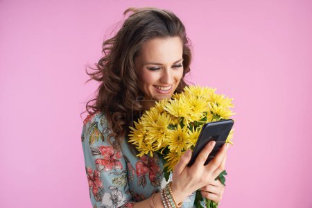 Foto de Mujer moderna feliz en vestido floral con flores de crisantemos amarillos enviando mensaje de texto con teléfono inteligente sobre fondo rosa. - Imagen libre de derechos