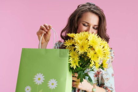 Photo for Happy trendy 40 years old woman with long wavy brunette hair with yellow chrysanthemums flowers and green shopping bag against pink background. - Royalty Free Image