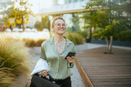 Téléchargez les photos : Femme d'affaires souriante moderne de 40 ans près du centre d'affaires en chemisier vert et lunettes avec mallette et écouteurs sans fil à l'aide d'un smartphone. - en image libre de droit