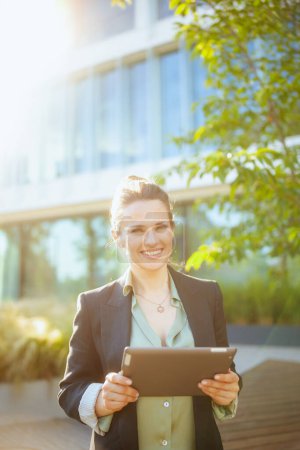 Téléchargez les photos : Portrait de travailleuse moderne souriante dans un quartier d'affaires en veste noire à l'aide d'une tablette PC. - en image libre de droit