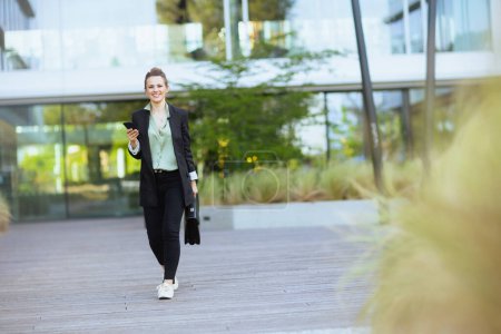 Foto de Retrato de larga duración de una trabajadora moderna sonriente en el distrito de negocios con chaqueta negra con maletín usando smartphone y caminando. - Imagen libre de derechos