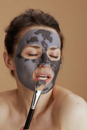 Foto de Joven mujer de 40 años con cepillo y máscara facial sobre fondo beige. - Imagen libre de derechos