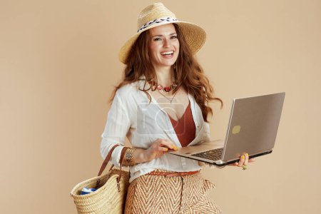 Téléchargez les photos : Vacances à la plage. heureuse élégante femme au foyer de 40 ans en chemisier blanc et short sur fond beige avec sac en paille, ordinateur portable et chapeau d'été. - en image libre de droit