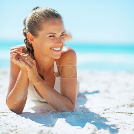 Téléchargez les photos : Jeune femme souriante allongée sur la plage et regardant au loin - en image libre de droit
