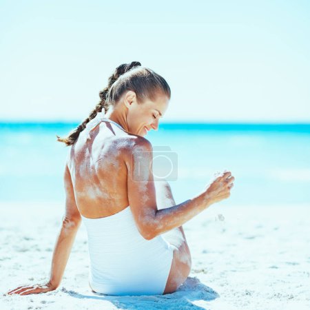 Foto de Mujer joven sonriente en traje de baño sentada en la playa y jugando con arena - Imagen libre de derechos