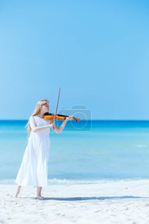 Foto de Retrato de larga duración de la adolescente moderna en vestido blanco en la orilla del océano con violín disfrutando jugando. - Imagen libre de derechos