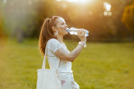 Foto de Hora de verano. mujer de 40 años de moda relajada en camisa blanca con bolsa de mano y botella de agua en el prado al aire libre. - Imagen libre de derechos
