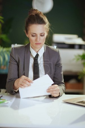 Photo for New job. pensive modern 40 years old woman employee in modern green office in grey business suit opening letter. - Royalty Free Image