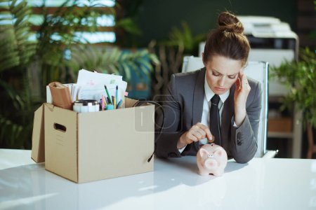 Photo for New job. stressed modern middle aged woman worker in modern green office in grey business suit with personal belongings in cardboard box putting coin into piggy bank. - Royalty Free Image