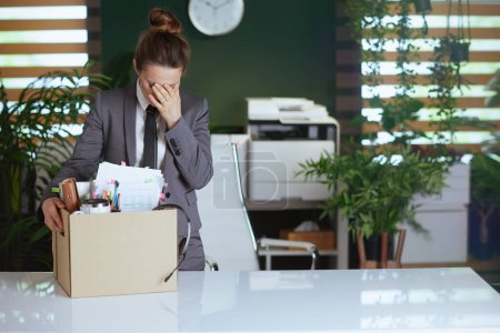 Un nuevo trabajo. triste mujer de mediana edad moderna trabajadora en moderna oficina verde en traje de negocios gris con pertenencias personales en caja de cartón.