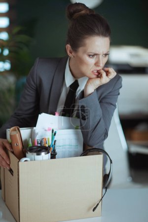 Photo for New job. concerned modern 40 years old woman worker in modern green office in grey business suit with personal belongings in cardboard box. - Royalty Free Image
