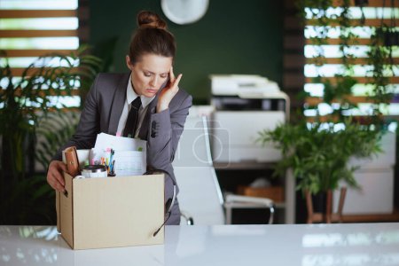 Un nuevo trabajo. trabajadora moderna preocupada en moderna oficina verde en traje de negocios gris con pertenencias personales en caja de cartón.