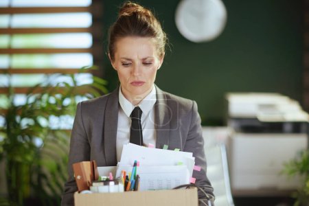 Un nuevo trabajo. trabajadora moderna infeliz en oficina verde moderna en traje de negocios gris con pertenencias personales en caja de cartón.
