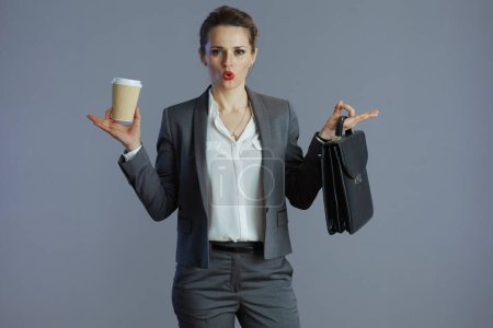 Téléchargez les photos : Jeune employée stressée en costume gris avec tasse à café et serviette isolée sur gris. - en image libre de droit