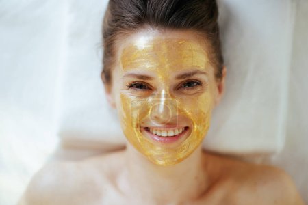 Foto de Tiempo de atención médica. Vista superior de la mujer moderna feliz en el salón de spa con máscara cosmética dorada en la cara que pone en la mesa de masaje. - Imagen libre de derechos