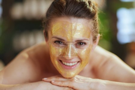 Foto de Tiempo de atención médica. sonriente mujer moderna de 40 años en el gabinete de masaje con máscara cosmética dorada en la cara puesta en la mesa de masaje. - Imagen libre de derechos