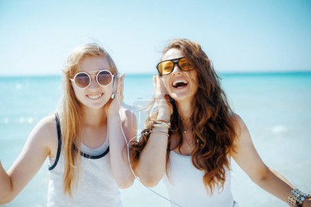 Téléchargez les photos : Mère moderne souriante et fille adolescente sur le littoral avec écouteurs. - en image libre de droit