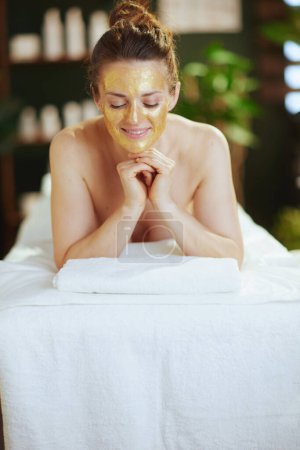 Foto de Tiempo de atención médica. mujer moderna relajada en el gabinete de masaje con máscara cosmética dorada en la cara que pone en la mesa de masaje. - Imagen libre de derechos