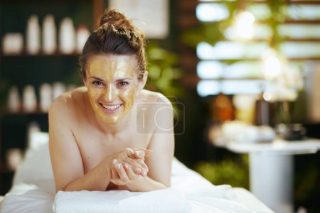 Foto de Tiempo de atención médica. mujer moderna sonriente en el gabinete de masaje con máscara cosmética dorada en la cara que pone en la mesa de masaje. - Imagen libre de derechos