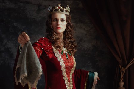 Téléchargez les photos : Reine médiévale en robe rouge avec mouchoir et couronne sur fond gris foncé. - en image libre de droit