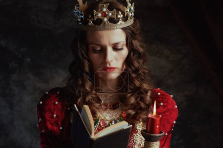 Téléchargez les photos : Reine médiévale en robe rouge avec livre, bougie et couronne sur fond gris foncé. - en image libre de droit
