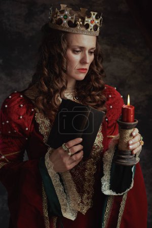 Photo for Medieval queen in red dress with book, candle and crown on dark gray background. - Royalty Free Image