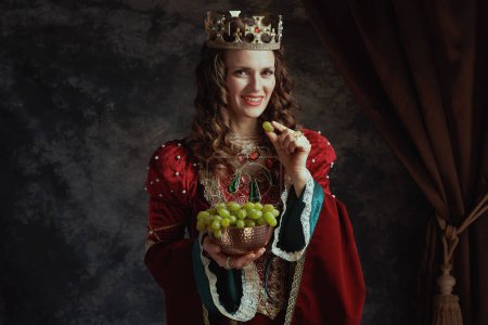 Foto de Feliz reina medieval en vestido rojo con plato de uvas y corona sobre fondo gris oscuro. - Imagen libre de derechos