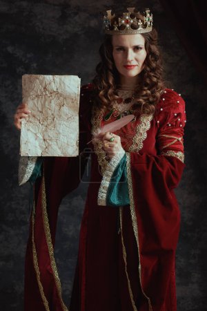 Photo for Happy medieval queen in red dress with parchment and crown on dark gray background. - Royalty Free Image