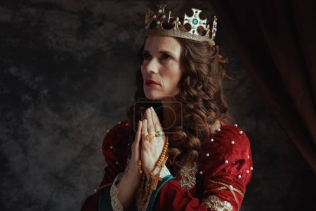mittelalterliche Königin in rotem Kleid mit Rosenkranz und Krone im Gebet auf dunkelgrauem Hintergrund.