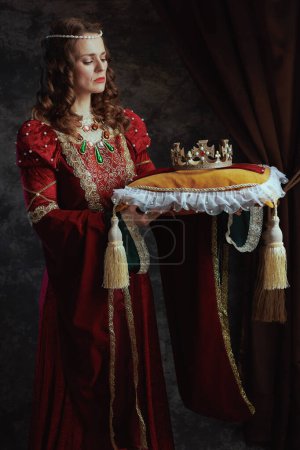 medieval queen in red dress with crown on pillow on dark gray background.