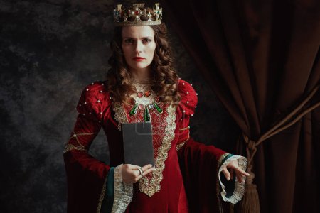 Photo for Medieval queen in red dress with book and crown on dark gray background. - Royalty Free Image