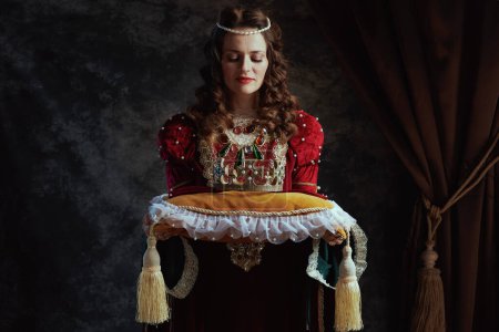 Foto de Reina medieval en vestido rojo con corona sobre almohada sobre fondo gris oscuro. - Imagen libre de derechos