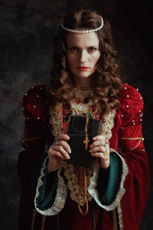 Foto de Reina medieval en vestido rojo con libro y rosario sobre fondo gris oscuro. - Imagen libre de derechos