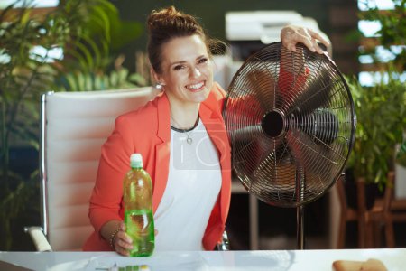 Photo for Sustainable workplace. Portrait of smiling modern small business owner woman at work in red jacket with bottle of water and electric fan. - Royalty Free Image
