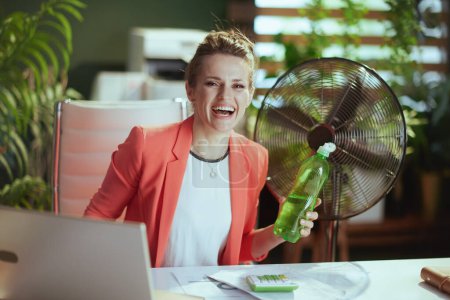 Téléchargez les photos : Un lieu de travail durable. Portrait d'heureuse femme d'affaires moderne de 40 ans au travail en veste rouge avec bouteille d'eau, ventilateur électrique et ordinateur portable. - en image libre de droit