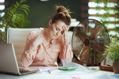 Photo for Sustainable workplace. modern small business owner woman in modern green office with electric fan and laptop suffering from summer heat. - Royalty Free Image