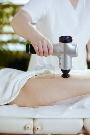 Temps de santé. Gros plan sur le massothérapeute médical dans le salon de spa avec pistolet de massage massant les clients jambe sur table de massage.