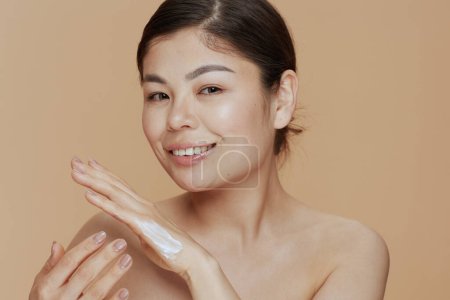 Téléchargez les photos : Portrait de femme asiatique moderne avec crème pour les mains isolée sur fond beige. - en image libre de droit