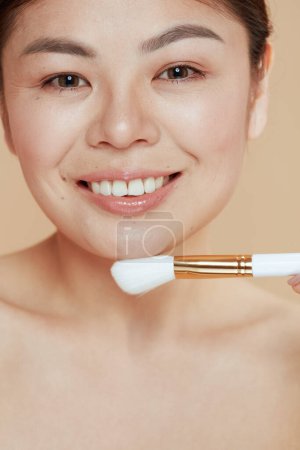 Téléchargez les photos : Portrait de femme asiatique moderne avec pinceau de maquillage sur fond beige. - en image libre de droit