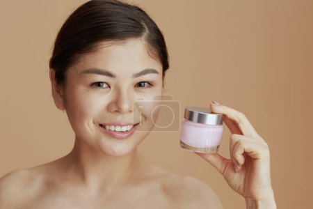 Foto de Retrato de joven hembra asiática con frasco de crema facial aislado sobre fondo beige. - Imagen libre de derechos