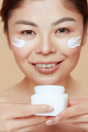 Foto de Retrato de mujer moderna con frasco de crema facial y crema facial en la cara aislada sobre fondo beige. - Imagen libre de derechos