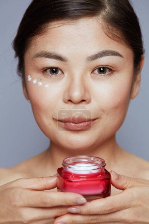 Foto de Moderna mujer asiática con crema facial tarro y crema facial en la cara aislado sobre fondo azul. - Imagen libre de derechos