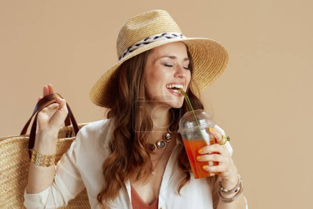 Photo for Beach vacation. happy modern middle aged housewife in white blouse and shorts isolated on beige with straw bag, carrot juice and summer hat. - Royalty Free Image