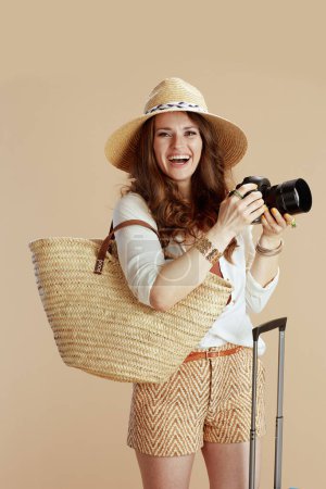 Photo for Beach vacation. smiling elegant middle aged housewife in white blouse and shorts isolated on beige with straw bag, trolley bag, photo camera and summer hat. - Royalty Free Image