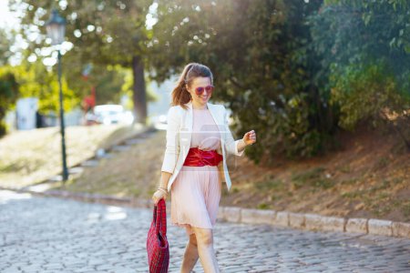 Photo for Happy trendy woman in pink dress and white jacket in the city with red bag and sunglasses walking. - Royalty Free Image