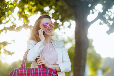 Téléchargez les photos : Heureuse jeune femme en robe rose et veste blanche dans la ville avec sac rouge et lunettes de soleil à l'aide d'un smartphone. - en image libre de droit