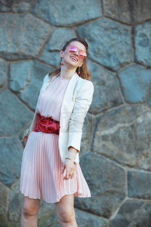 Téléchargez les photos : Souriant élégant femme de 40 ans en robe rose et veste blanche dans la ville avec des lunettes de soleil contre le mur de pierre. - en image libre de droit