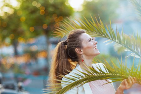 Téléchargez les photos : Femme souriante moderne de 40 ans en robe rose et veste blanche dans la ville près d'un arbre tropical. - en image libre de droit