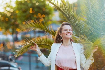 Téléchargez les photos : Femme élégante souriante en robe rose et veste blanche dans la ville près de l'arbre tropical. - en image libre de droit