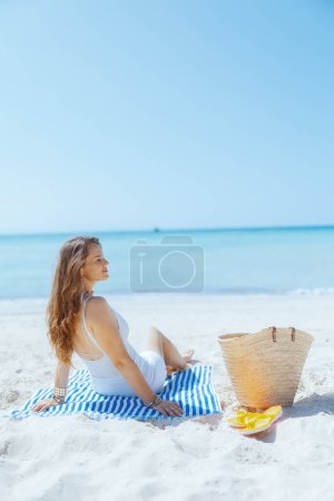 Téléchargez les photos : Femme moderne détendue de 40 ans sur le bord de l'océan avec sac de paille et serviette rayée. - en image libre de droit